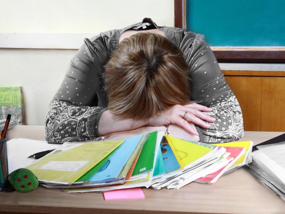 Teacher with head on desk on top of a pile of folders.