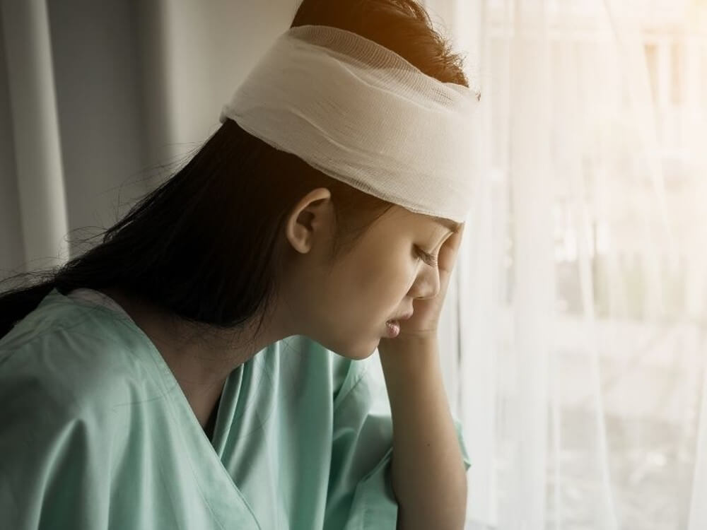 Woman with a bandaged head leans against a window.