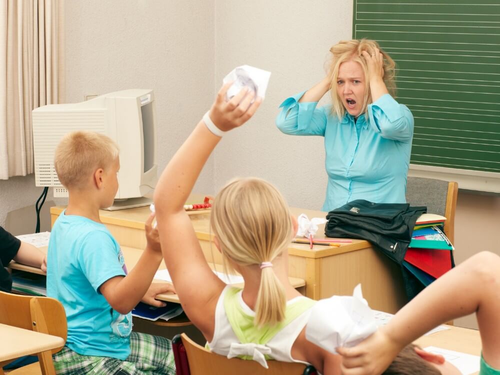Teacher looking frustrated while children throw wads of paper.